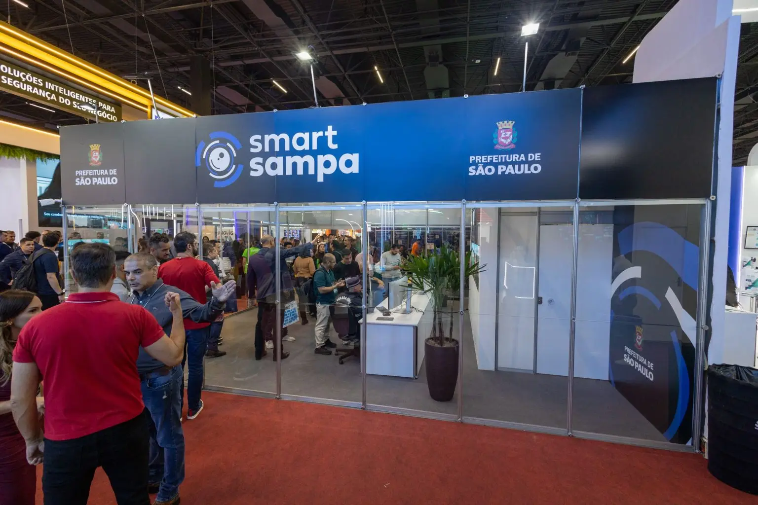 Profissionais de segurança e visitantes interagem com painel tecnológico do projeto Smart Sampa durante exposição de tecnologia de segurança.